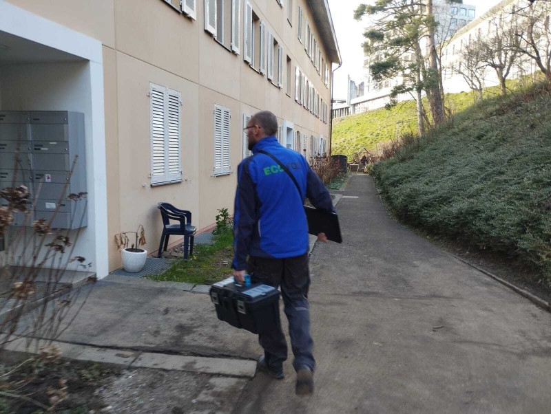 Un technicien en lutte antiparasitaire marche dans une all&eacute;e du canton de Vaud, portant une mallette d&rsquo;intervention et se rendant vers un b&acirc;timent r&eacute;sidentiel.